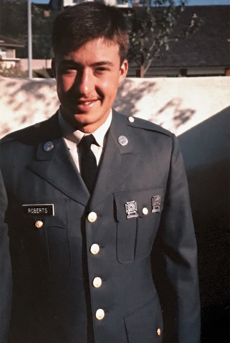 A young Gary Roberts smiling in his military dress uniform with a ROBERTS nameplate and marksmanship badges, photographed outdoors on a sunny day.