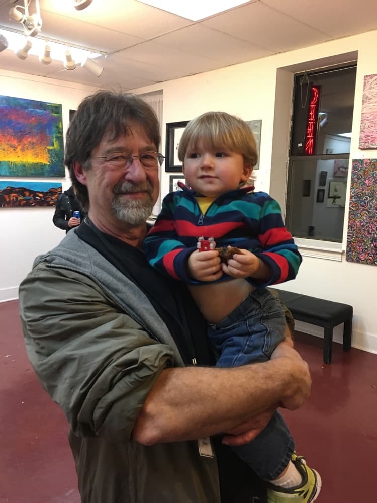 Gary Roberts smiling and holding a young boy in a striped jacket inside Soulard Art Gallery, with paintings on the walls behind them.