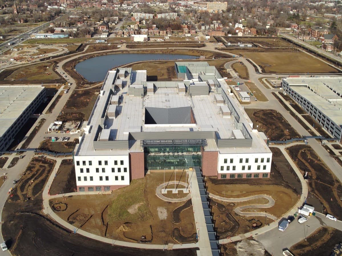 Aerial view of the National Geospatial-Intelligence Agency's new St. Louis campus under construction, with a large modern headquarters building, parking structures, a lake, and the city skyline in the distance.